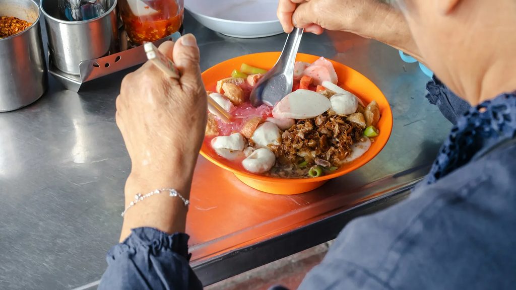 A diner eating a bowl of pink yen ta fo noodles at a stainless steel street food counter in Bangkok, the kind of Thai culture and Bangkok street food that anchors the Amazing Thailand Food Festival