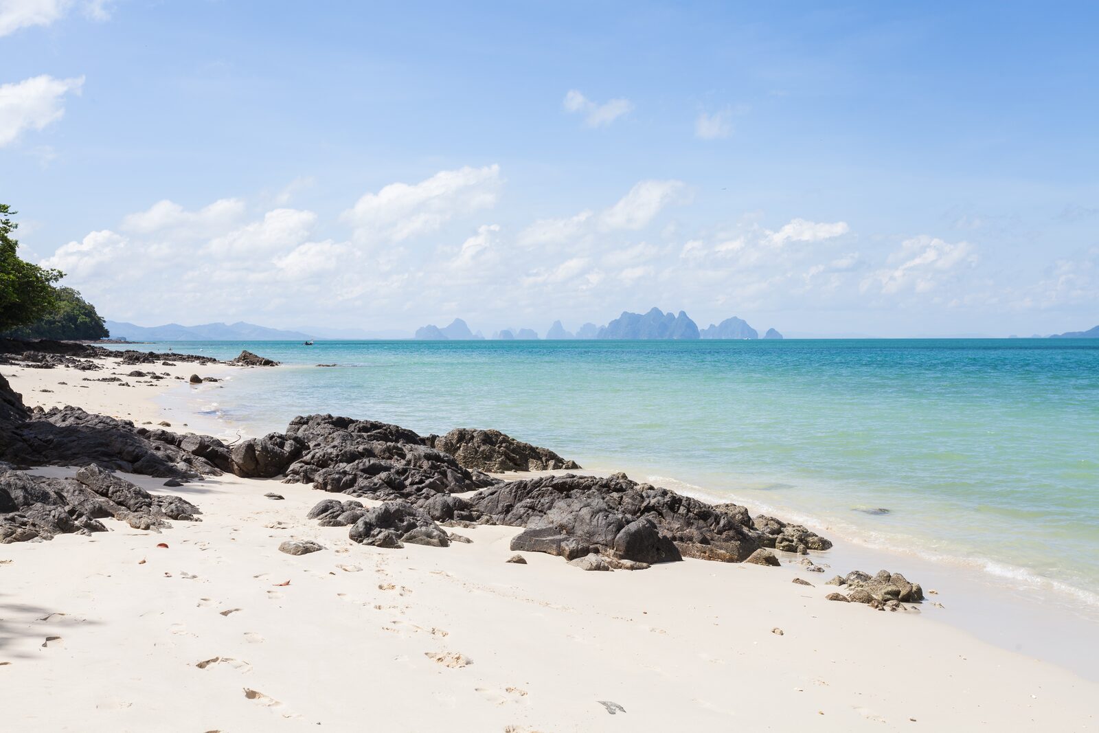 A quiet white-sand beach with turquoise water and limestone karsts on the horizon off Phuket, typical of the Thailand islands reachable on a short boat trip
