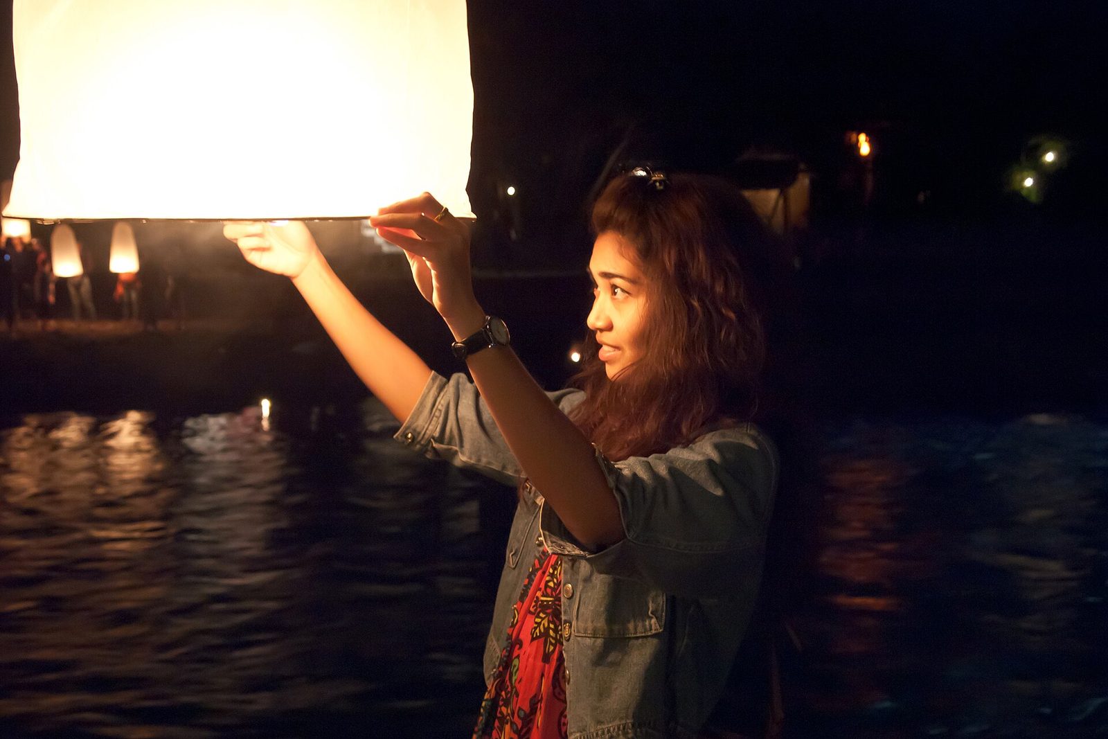 A woman releasing a glowing paper sky lantern beside a dark river at night during Loy Krathong in Chiang Mai, the free riverside version of Thai festivals 2026