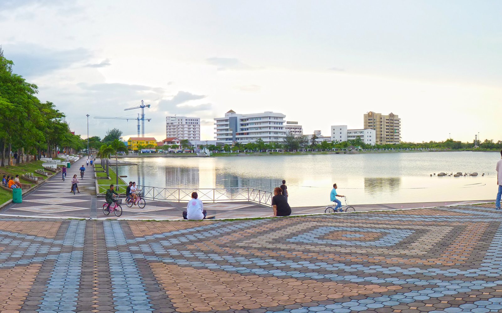 Cyclists and families on the patterned lakeside promenade of Nong Prajak Park in Udon Thani at dusk, the city park at the centre of a northeastern Thailand itinerary