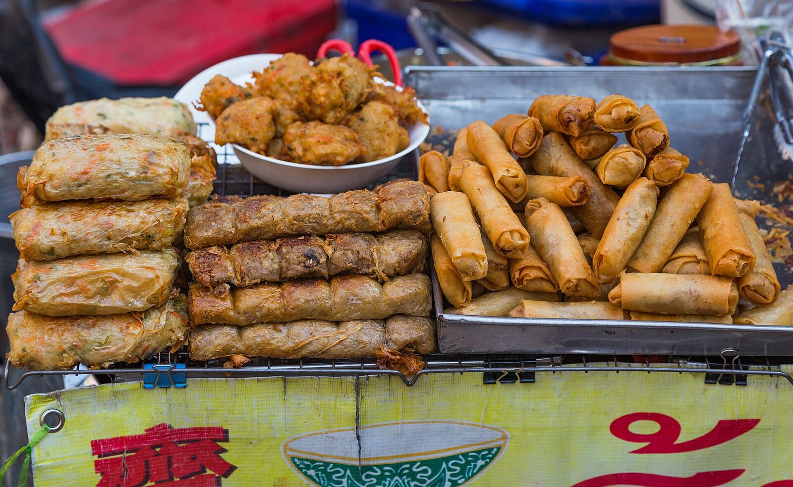 A Yaowarat street food cart in Bangkok's Chinatown piled with fresh and fried spring rolls, fish cakes and pork rolls on wire racks, the kind of stall the planned Food Express layover tour is built around