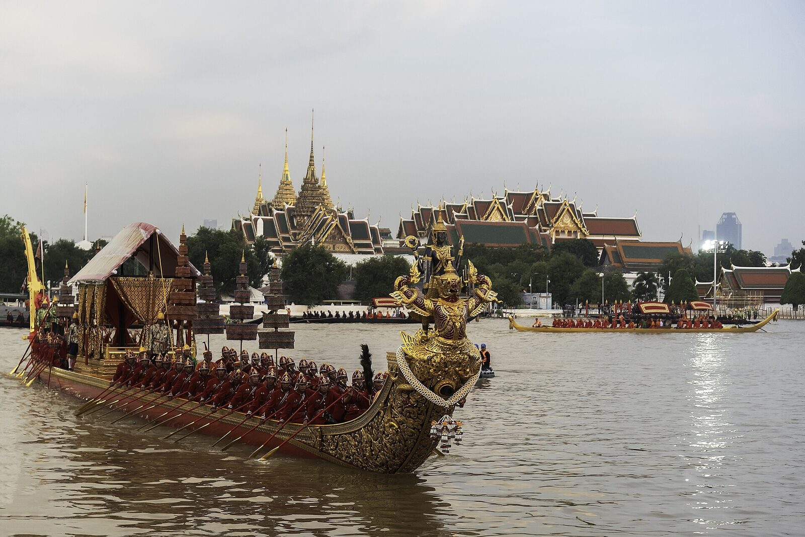 The gilded royal barge Narai Song Suban with rows of red-uniformed Royal Thai Navy oarsmen passing the Grand Palace on the Chao Phraya River during a Royal Barge Procession rehearsal, a free Bangkok tourism spectacle in October 2026