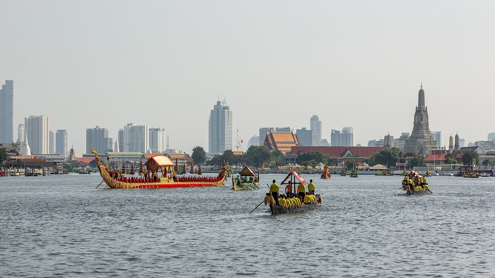 Gilded Thai royal barges in formation on the Chao Phraya River during a Royal Barge Procession rehearsal, with the Bangkok skyline and the prang of Wat Arun behind