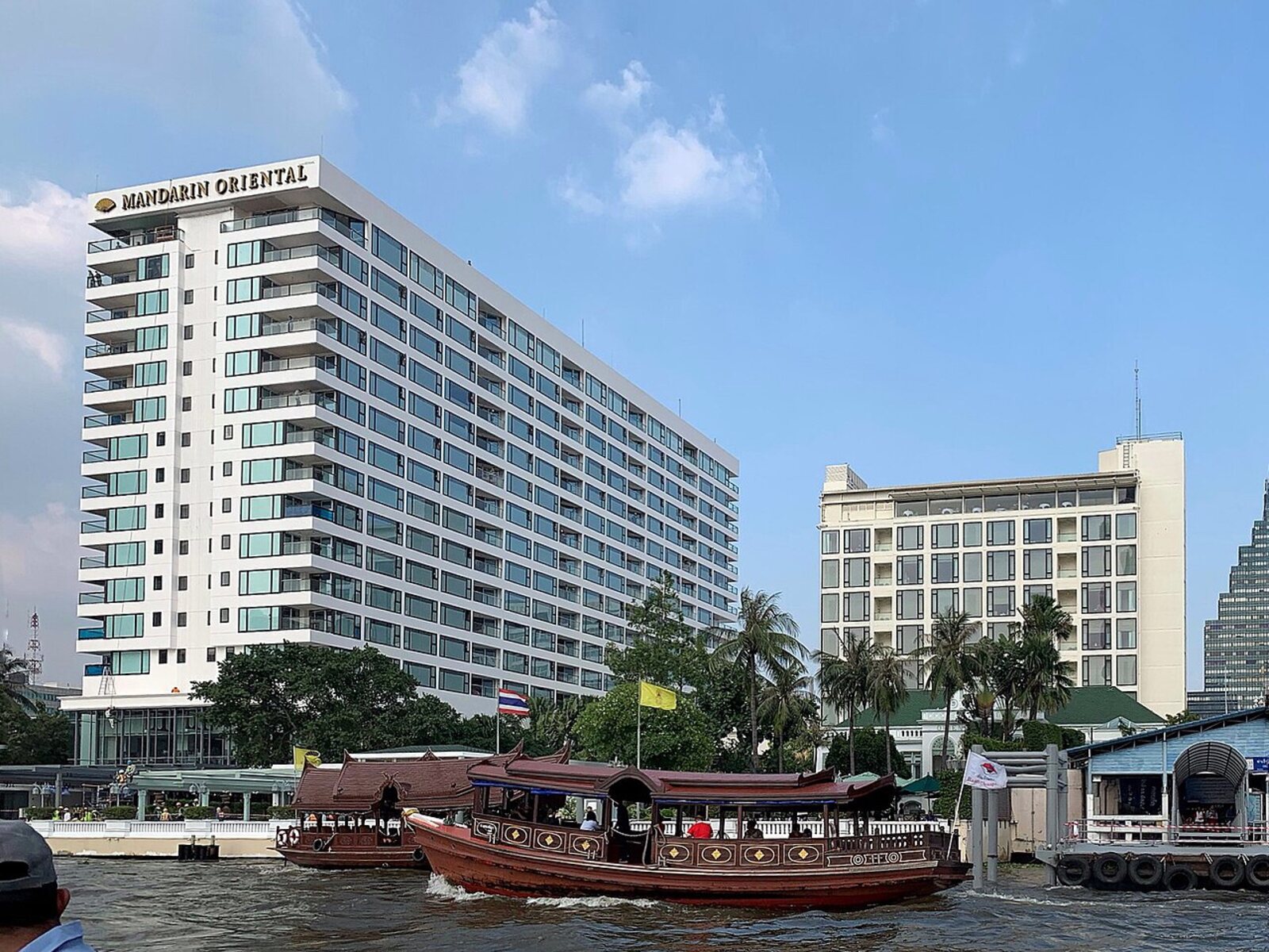 The riverside facade of the Mandarin Oriental Bangkok on the Chao Phraya with a teak shuttle boat passing, voted the best hotel in Southeast Asia in the 2026 Readers' Choice Awards