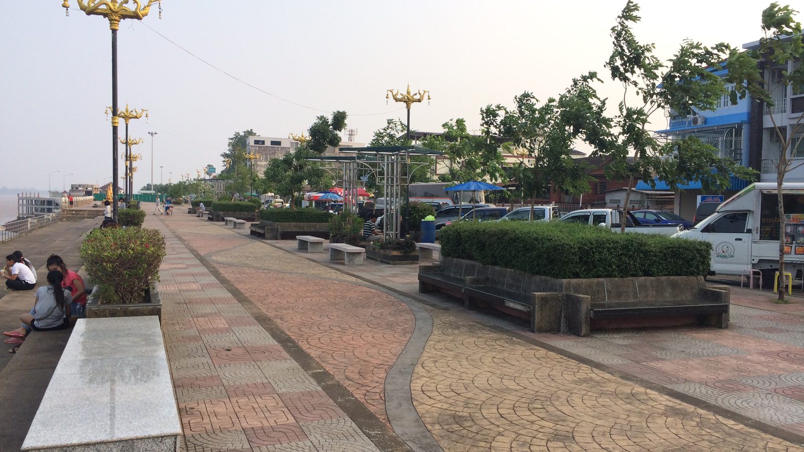 The Mekong riverfront promenade at Nakhon Phanom, lined with gilded naga lamp posts and stone benches, where the illuminated boat procession is watched during Thai festivals 2026