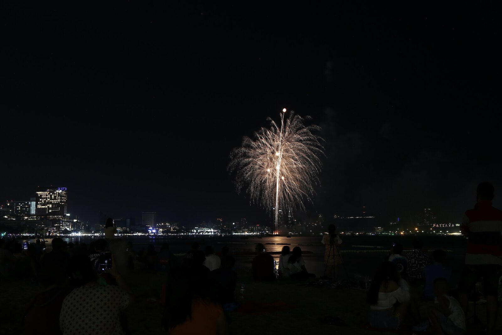 A golden firework bursting over Pattaya Bay at the Pattaya International Fireworks Festival, watched by crowds sitting on the sand with the illuminated PATTAYA hillside sign on the right