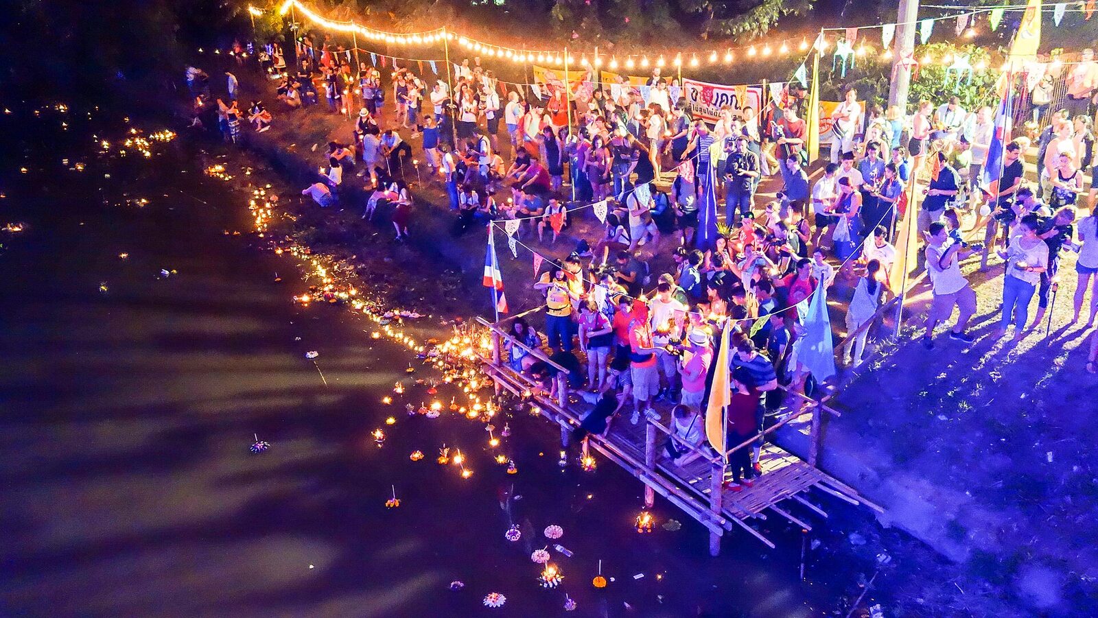 Crowds floating candle-lit banana-leaf krathongs from a bamboo jetty at night during Loy Krathong in Thailand, with lights strung above the riverbank, one of the biggest Thai festivals of 2026
