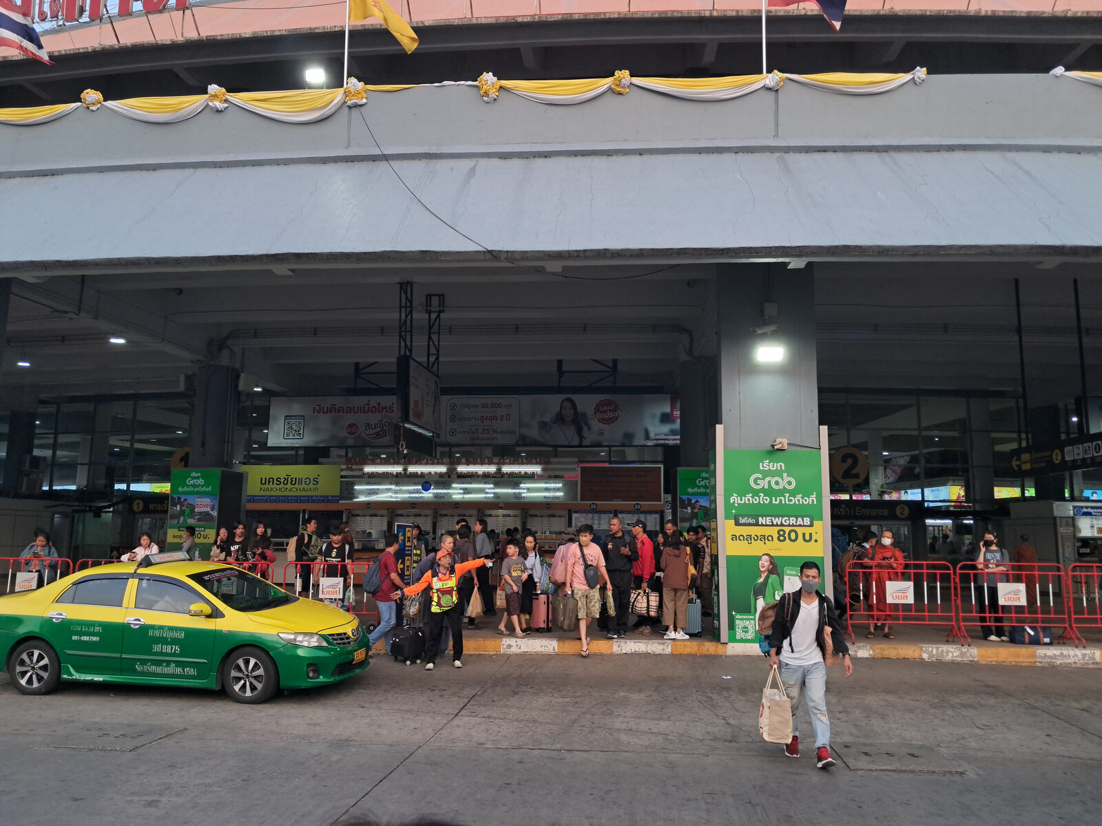Travellers with suitcases outside the ticket counters at Mo Chit 2 Bus Terminal in Bangkok, with a green and yellow taxi waiting at the kerb, the main departure point for long-weekend trips on a Thailand itinerary