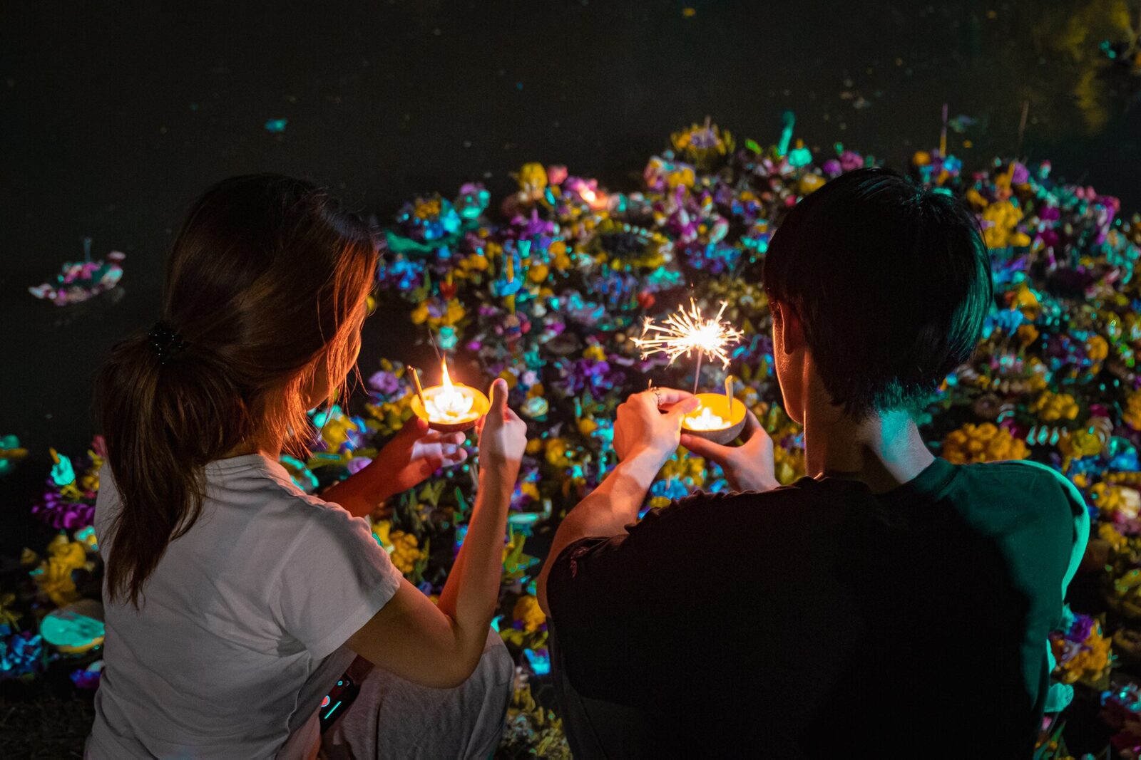 Two people kneeling at the water's edge holding lit krathongs with a candle and a sparkler as hundreds of flower floats drift past, the central ritual of Loy Krathong and of Thai culture