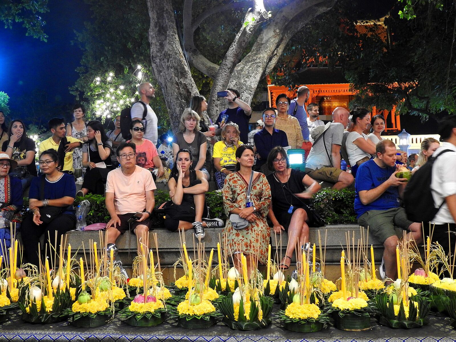 Rows of banana-leaf krathongs with yellow candles lined up for sale in front of a seated festival crowd on Loy Krathong night, a scene to budget for in any Thailand itinerary