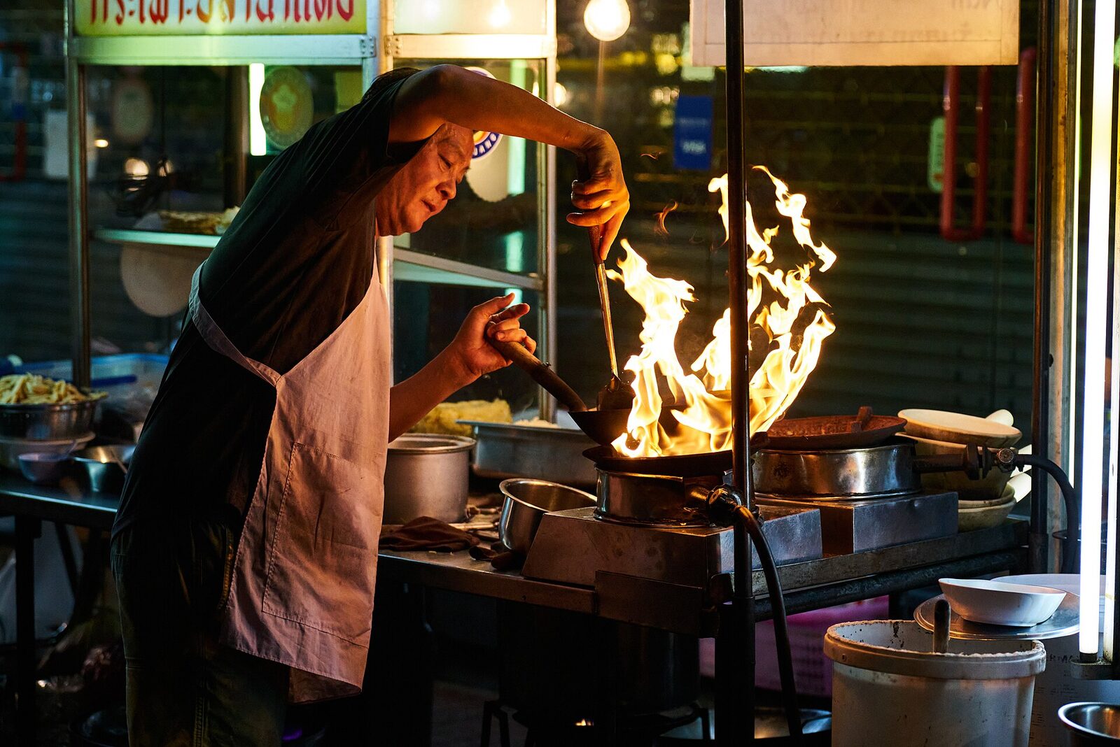 A Bangkok street food cook leaning over a flaming wok at a night stall in Chinatown, the kind of evening a saved 1,000 baht buys on a Thailand itinerary
