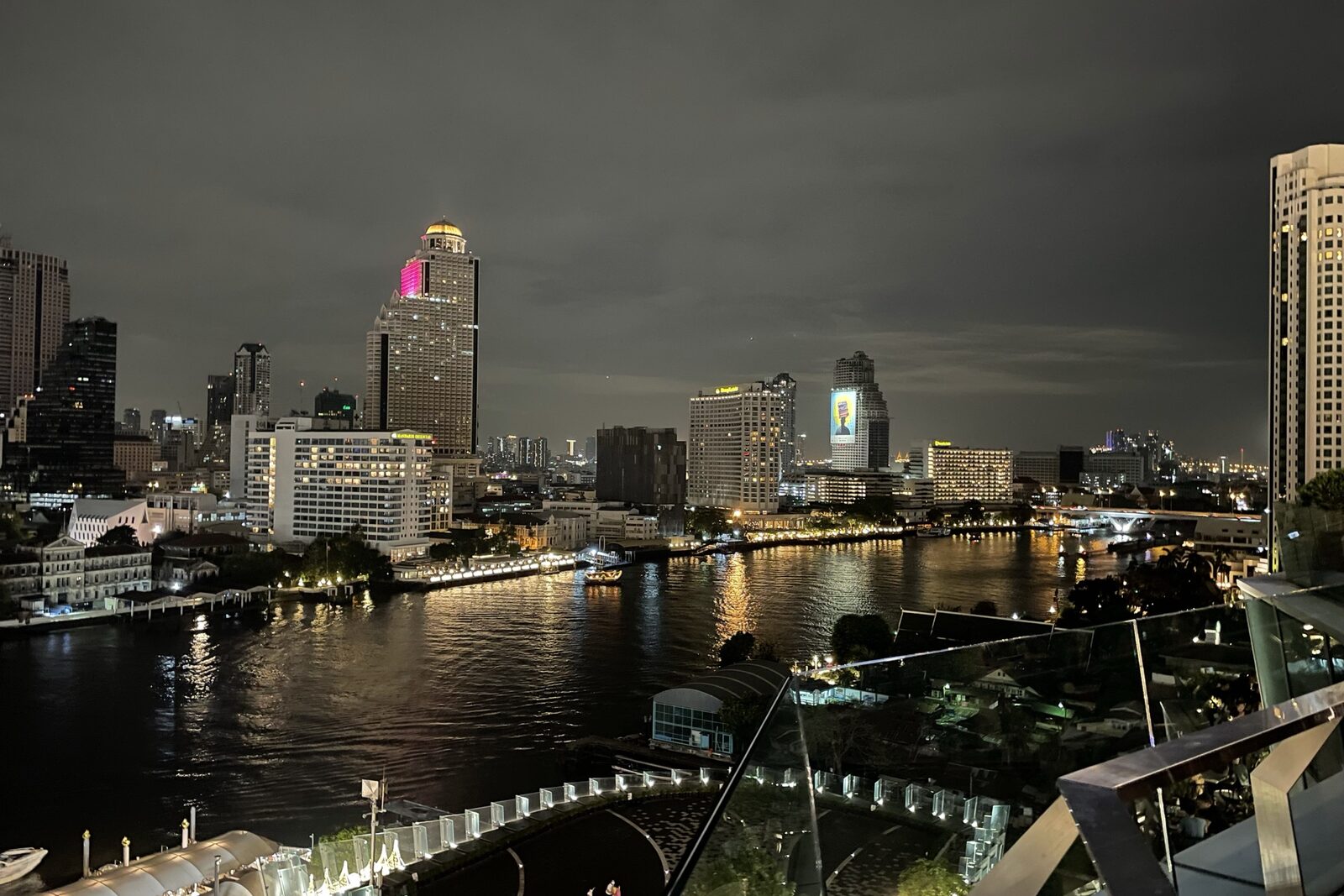 The Chao Phraya River at night in Bangkok with lit hotel towers, riverside piers and a passing boat trailing reflections, the setting for the Vijit Chao Phraya light festival and a Bangkok tourism highlight in Thailand