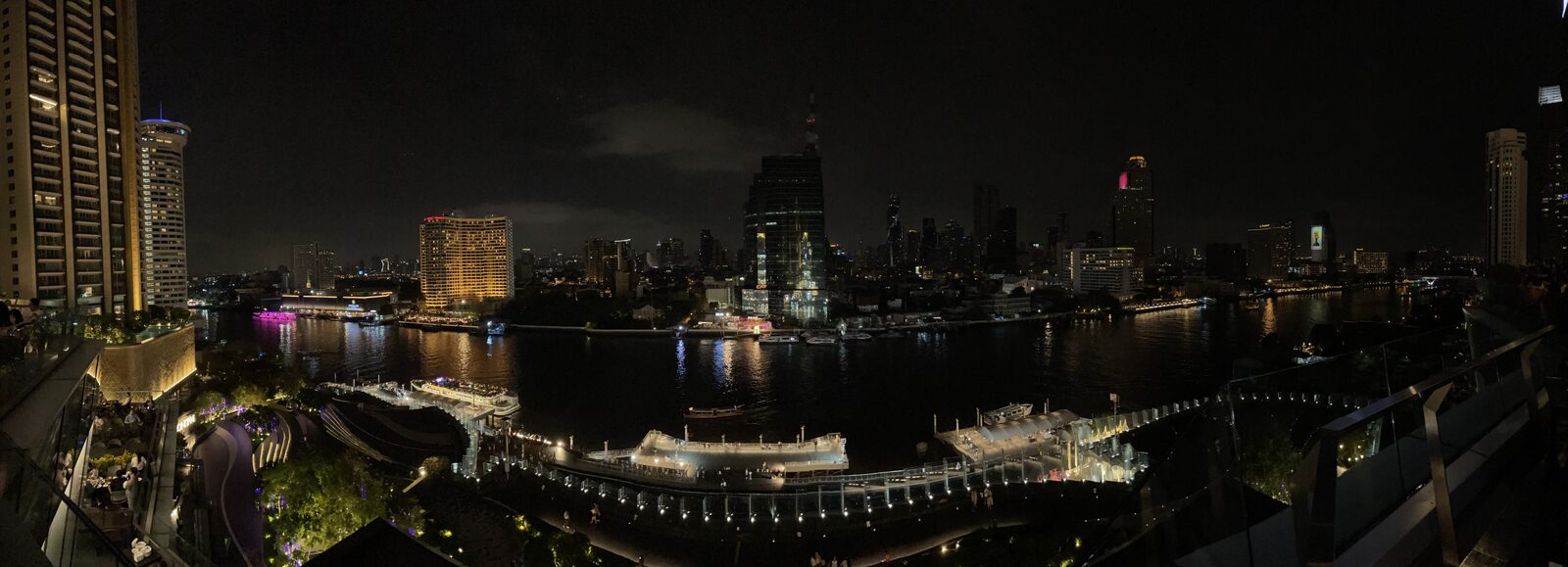 Wide night panorama of the Chao Phraya River in Bangkok with the lit riverside boardwalk, moored boats and the city skyline, the backdrop to Vijit Chao Phraya and a classic stop on a Thailand itinerary