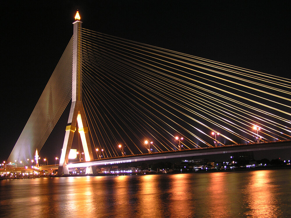 The harp-shaped Rama VIII Bridge in Bangkok lit gold at night with its cables reflected in the Chao Phraya River, one of the 13 Vijit Chao Phraya festival sites and a classic Bangkok tourism view