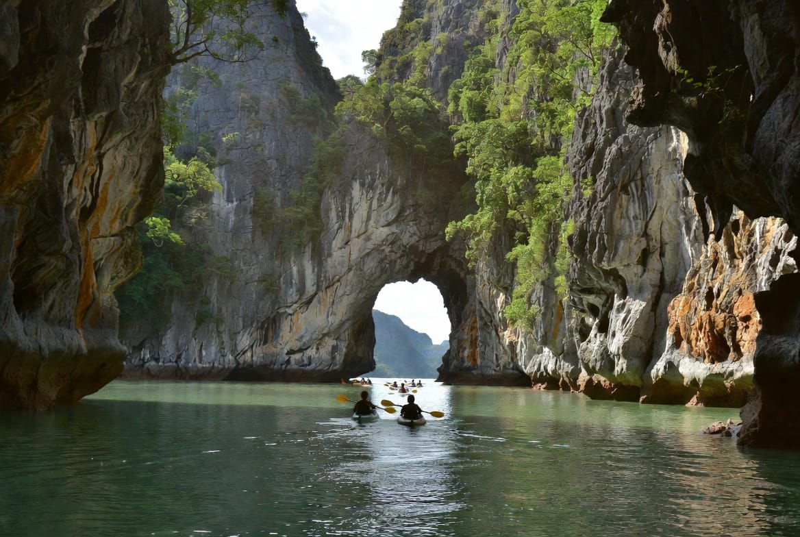 Kayakers paddling through a limestone cave arch in Phang Nga Bay
