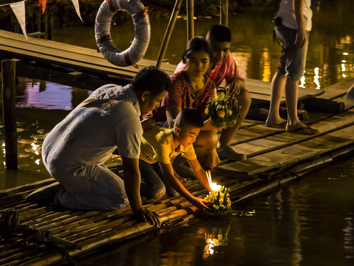 People crouching at the river's edge to set candle-lit krathongs onto the Ping river at night