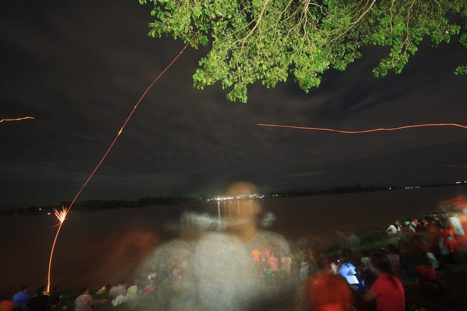 Crowds on the Mekong riverbank at night watching light trails rise over the water during the Naga Fireballs festival